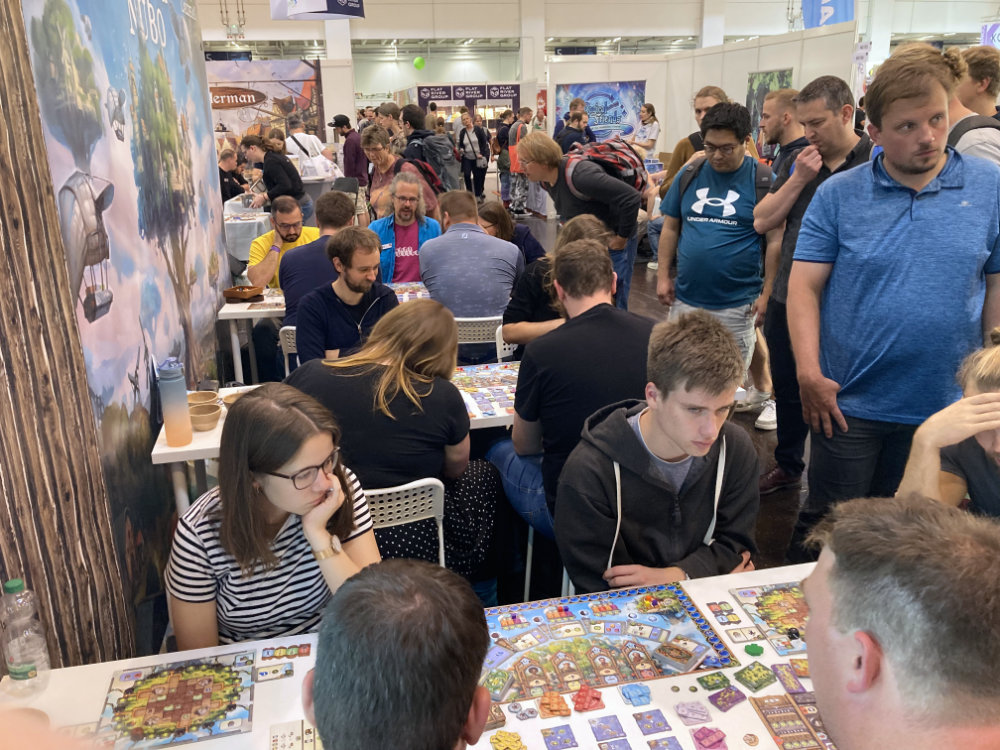 Blick auf die drei voll besetzten Planta-Nubo-Spieltische am The-Game-Builders-Stand auf der Spiel ´23. Drumherum stehen noch viele wartende Personen. Am letzten Spieltisch erklärt der Autor Mike Keller in blauer Jacke das Spiel.