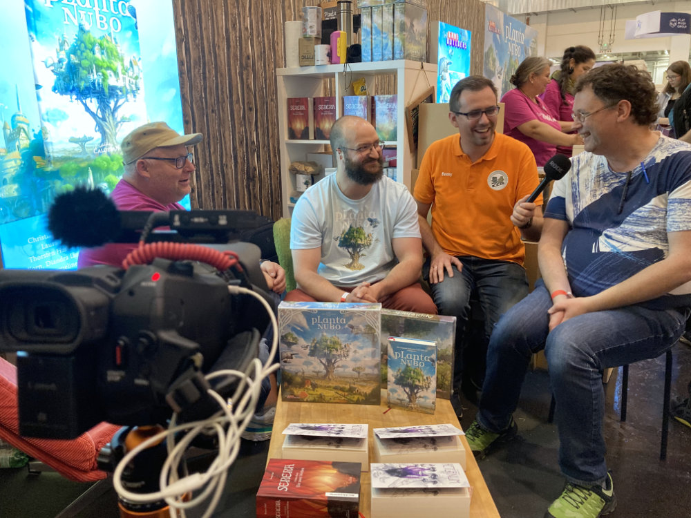Am Gamebuilders-Stand um einen Tisch sitzend, im Vordergrund filmt eine Kamera das Gespräch mit: ode., Lukas Siegmon, Benny von den BrettspielSuchties mit einem Mikrofon in der Hand, Uwe Rosenberg, der gerade eine Frage beantwortet.