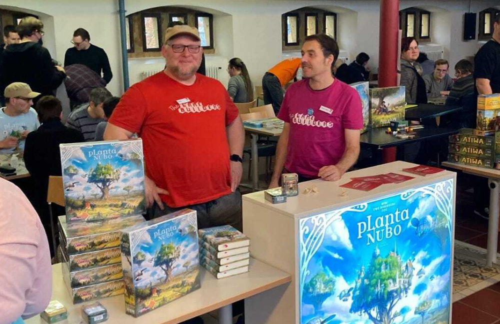 ode. und Thorsten hinter einer beleuchteten Theke mit Planta-Nubo-Motiv vor Spieletischen mit Spieler*innen im Historischen Saal der Ravensberger Spinnerei in Bielefeld